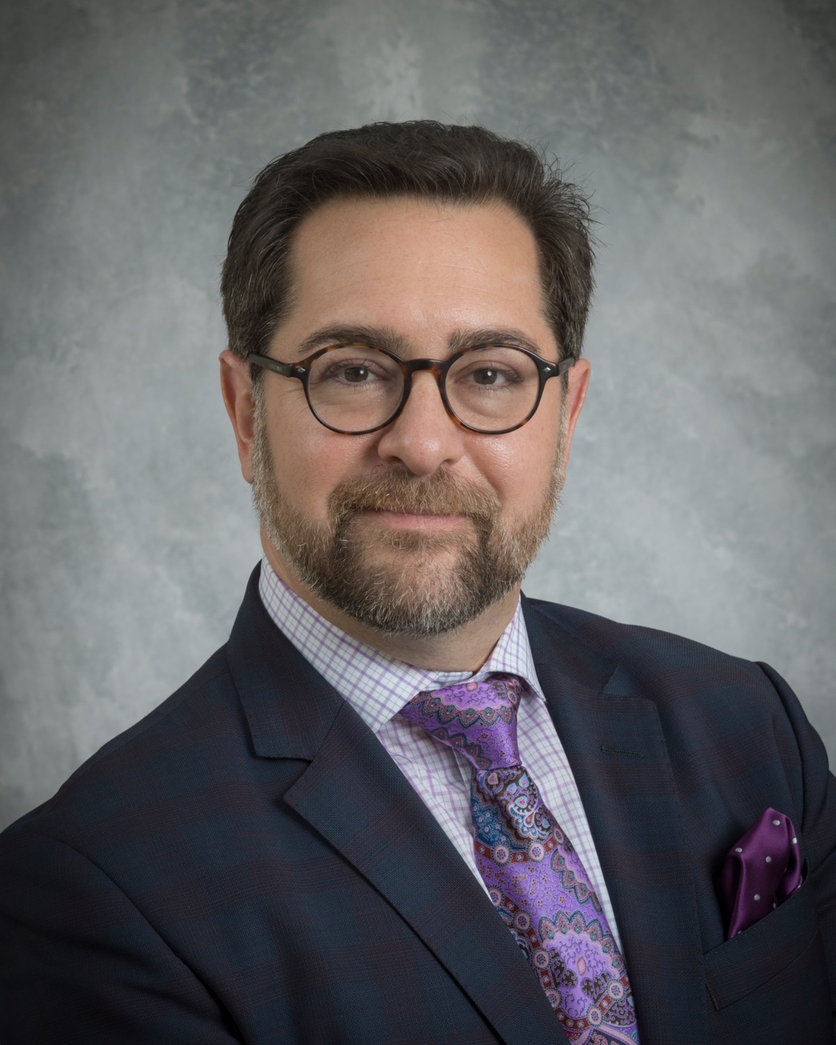 Portrait of Gregory Hansell in a navy suit with a richly patterned purple and blue tie